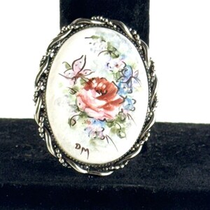 May include: An oval-shaped brooch with a silver-toned twisted wire frame. The center of the brooch features a white porcelain cameo with a hand-painted floral design in shades of pink, blue, and green. The cameo also includes a butterfly and the initials "DM".