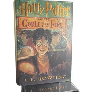 Puede incluir: Un libro de tapa dura titulado "Harry Potter y el cáliz de fuego" de J.K. Rowling. La portada presenta una ilustración de Harry Potter con otros personajes y un fondo de fuego. El libro se exhibe sobre un soporte de acrílico transparente.