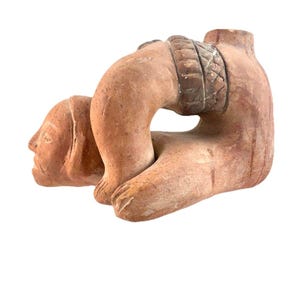 Pode incluir: Escultura em terracota que retrata uma figura ajoelhada. A figura tem cabeça, braços e pernas, com um cinto detalhado em volta da cintura. A escultura tem um tom quente e terroso, com textura e desgaste visíveis.