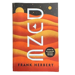 May include: A paperback book titled "DUNE" by Frank Herbert. The cover features a desert landscape in shades of orange and yellow, with the title in white and a small figure walking across the dunes. Text on the cover reads "SOON TO BE A MAJOR MOTION PICTURE."
