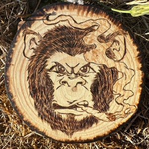 May include: A wood-burned image of a monkey smoking a cigarette on a wooden disc. The monkey has a serious expression and is surrounded by smoke.