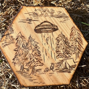 May include: Hexagon-shaped wooden art piece with a pyrography design. The artwork depicts a UFO abducting a figure above a campsite with trees and a campfire. The wood has a natural, light brown color with a darker, burnt edge.