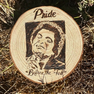 May include: A wood-burned image of a man with the text "Pride Before the Fall" on a wooden round.