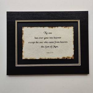 May include: A framed print with a black background and a gold border. The print features a quote from the Bible, John 3:13, written in a cursive font on a parchment-like paper. The quote reads: "No one has ever gone into heaven except the one who came from heaven - the Son of Man."