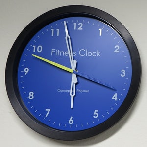 May include: A round wall clock with a black frame and a blue face. The clock face features white numerals, white hands, and the text "Fitness Clock." The minute hand is yellow, and the second hand is blue. The text "Concept Polymer" is also visible.
