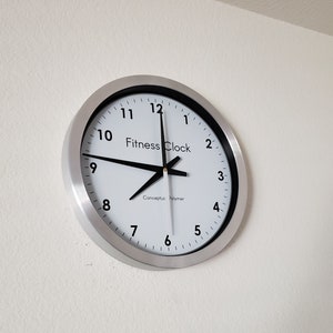 May include: A round wall clock with a silver-colored metal frame. The clock face is white with black numerals and hands. The text "Fitness Clock" is printed on the face. The clock is mounted on a white wall.