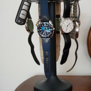 May include: A black watch stand displaying several wristwatches with leather and fabric straps. A car key fob hangs on the left. The watches have silver, black, and blue faces. The stand is black with a round base.