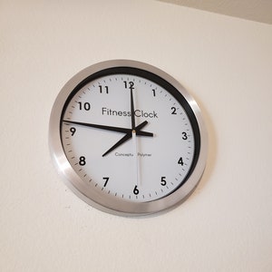 May include: A round wall clock with a silver-colored metal frame. The clock face is white with black numerals and hands. The text "Fitness Clock" is printed on the face. The time displayed is approximately 8:00.