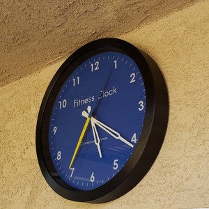 May include: A round black wall clock with a blue face. The clock has white numerals and hands, with a yellow second hand. The words "Fitness Clock" are printed on the face. The clock is mounted on a textured, tan wall.