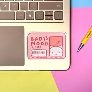 May include: A pink sticker with the text "BAD MOOD CLUB" and a cartoon cat illustration is placed on a laptop. The sticker also includes a barcode, the words "LIFETIME MEMBER" and "ID-XXX-XXX". A yellow pen with the text "THIS MEETING" is also visible.