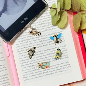 May include: A Kindle e-reader rests on an open book with a pink pen. Several insect-themed enamel pins, including a scorpion, bee, butterfly, and dragonfly, are displayed on the pages. The book has the title "Agustina Bazterrica, A Novel."