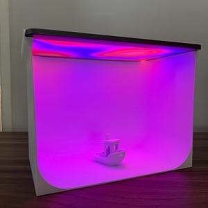 Peut inclure: Un petit bateau jouet blanc se trouve au centre d'un studio photo blanc en forme de cube avec une source de lumière rose et violette.