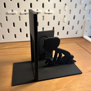 K&ouml;nnte beinhalten: Schwarze, 3D-gedruckte Buchst&uuml;tzen mit einer Silhouette einer knienden Person. Die Buchst&uuml;tzen haben eine rechteckige Basis und eine vertikale St&uuml;tze. Das Design ist minimalistisch und modern, ideal f&uuml;r B&uuml;cher.