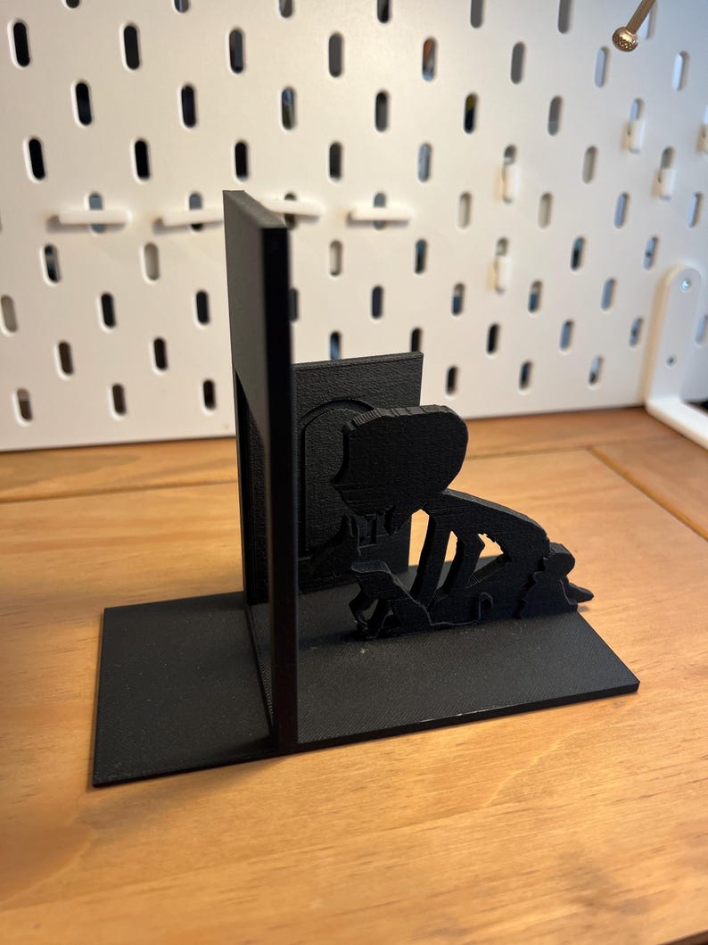 K&ouml;nnte beinhalten: Schwarze, 3D-gedruckte Buchst&uuml;tzen mit einer Silhouette einer knienden Person. Die Buchst&uuml;tzen haben eine rechteckige Basis und eine vertikale St&uuml;tze. Das Design ist minimalistisch und modern, ideal f&uuml;r B&uuml;cher.