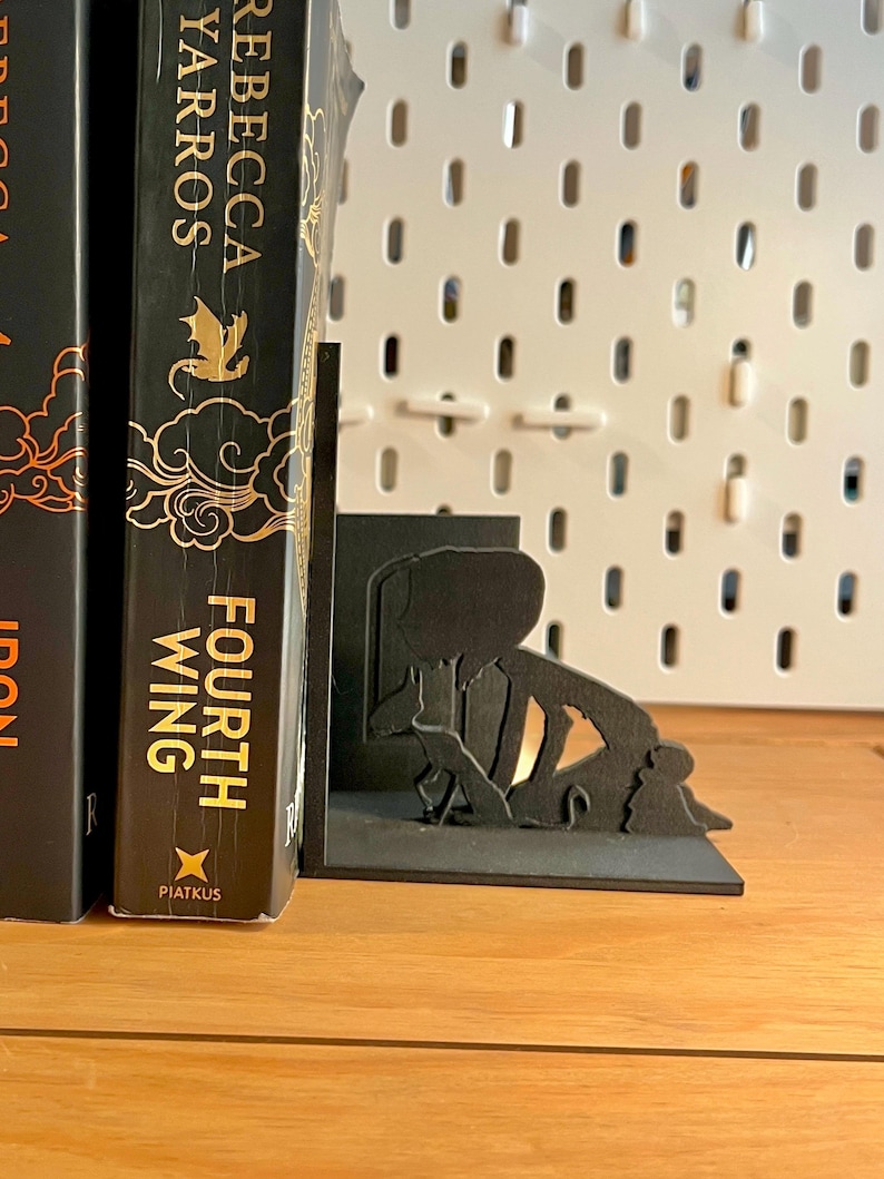 K&ouml;nnte beinhalten: Schwarze Metall-Buchst&uuml;tzen mit einer Silhouette einer lesenden Person. Die Buchst&uuml;tzen halten ein Buch mit dem Titel "Fourth Wing" von Rebecca Yarros. Der Hintergrund ist eine wei&szlig;e Lochwand.