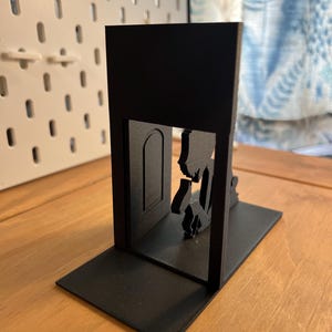 K&ouml;nnte beinhalten: Ein schwarzer, 3D-gedruckter Buchst&uuml;tze mit einer Silhouette einer Person, die durch eine T&uuml;r geht. Die Buchst&uuml;tze hat eine rechteckige Basis und einen vertikalen Abschnitt mit dem T&uuml;rdesign. Das Design ist modern und minimalistisch.