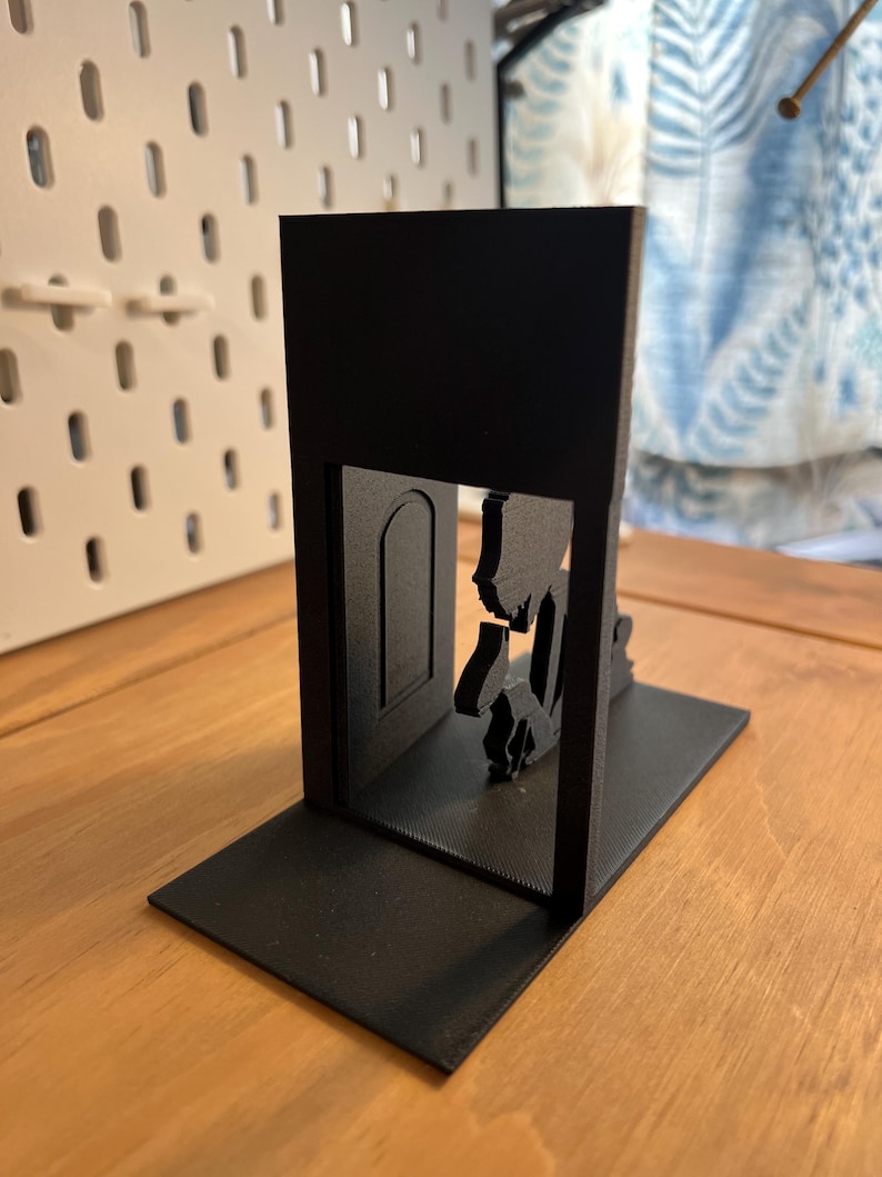 K&ouml;nnte beinhalten: Ein schwarzer, 3D-gedruckter Buchst&uuml;tze mit einer Silhouette einer Person, die durch eine T&uuml;r geht. Die Buchst&uuml;tze hat eine rechteckige Basis und einen vertikalen Abschnitt mit dem T&uuml;rdesign. Das Design ist modern und minimalistisch.