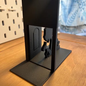 K&ouml;nnte beinhalten: Schwarzer Metall-Buchst&uuml;tze mit einer Silhouette eines T&uuml;rrahmens, durch den eine Figur geht. Die Buchst&uuml;tze hat eine rechteckige Basis und eine vertikale St&uuml;tze mit dem T&uuml;rausschnitt. Das Design ist minimalistisch und modern.