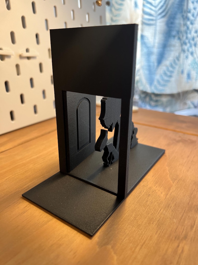 K&ouml;nnte beinhalten: Schwarzer Metall-Buchst&uuml;tze mit einer Silhouette eines T&uuml;rrahmens, durch den eine Figur geht. Die Buchst&uuml;tze hat eine rechteckige Basis und eine vertikale St&uuml;tze mit dem T&uuml;rausschnitt. Das Design ist minimalistisch und modern.