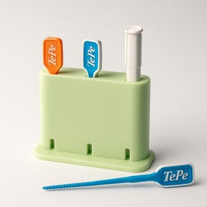 Puede incluir: Un organizador de higiene dental verde claro con tres ranuras, que contiene artículos naranjas, azules y blancos con la etiqueta "TePe". Un palillo dental azul con el logotipo "TePe" descansa al frente. El organizador está sobre un fondo blanco.