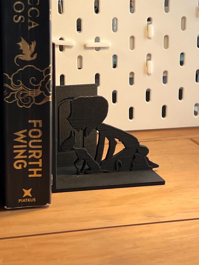 K&ouml;nnte beinhalten: Schwarze Silhouette eines Buchst&uuml;tze mit einer Person, zwei Hunden und einer Katze. Die Buchst&uuml;tze steht auf einer Holzoberfl&auml;che, mit einem Buch auf der linken Seite. Das Buch tr&auml;gt den Titel "Fourth Wing".