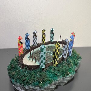 Może przedstawiać: Miniaturowy model boiska do Quidditcha. Model przedstawia okrągłe boisko ze słupkami do bramek i wieżami w szachownicę w różnych kolorach. Podstawa to kamienista konstrukcja pokryta zielonym mchem.
