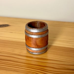 May include: A small wooden barrel with silver metal bands. The barrel is brown and has a wood grain texture.