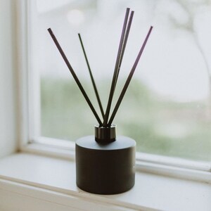 May include: A black essential oil diffuser with dark reeds. The cylindrical container is on a white windowsill, with a blurred green background visible through the window. The reeds are arranged in a fan shape.