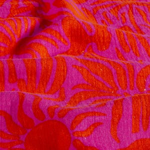 Gros plan sur un textile vibrant avec un motif répété de feuilles de palmier orange et un soleil rouge sur un fond rose vif. Le tissu est doux et texturé, avec des plis visibles et de subtiles variations de couleur.