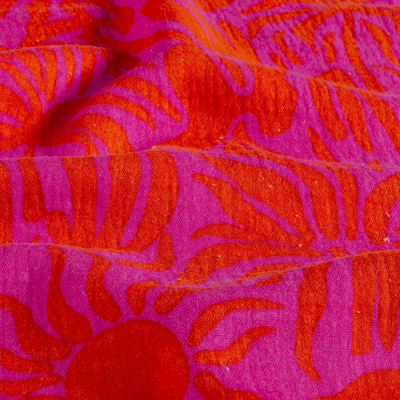 Gros plan sur un textile vibrant avec un motif répété de feuilles de palmier orange et un soleil rouge sur un fond rose vif. Le tissu est doux et texturé, avec des plis visibles et de subtiles variations de couleur.