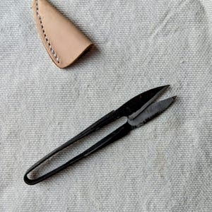 Può includere: Forbici da ricamo in metallo nero con lama curva e manici ad anello, insieme a un fodero in pelle marrone chiaro. Le forbici sono progettate per il taglio di precisione nei lavori di cucito e nei progetti di artigianato.