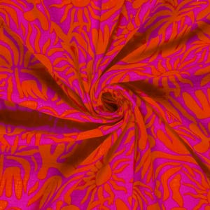 Tombé d'un tissu en gaze de coton rose vif avec un motif répété de formes orangées en forme de soleil. Le tissu est rassemblé en spirale son tombé est parfait pour des vêtements et accessoires d'été