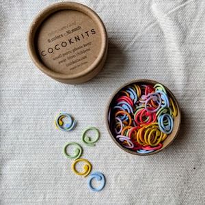 Może przedstawiać: Małe kartonowe pudełko z pokrywką, wypełnione kolorowymi plastikowymi znacznikami ściegów. Pudełko ma nadrukowany tekst "nylon coated steel 6 colors - 10 each COCOKNITS small parts, please keep away from children cocoknits.com made in china".