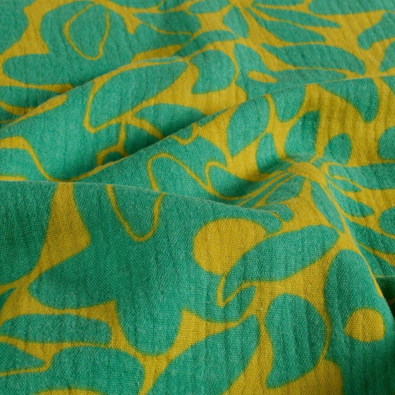 May include: Close-up of a fabric with a repeating floral pattern. The design features large, stylized leaves in a vibrant yellow against a teal green background. The fabric has a soft, textured appearance, with visible folds and shadows.