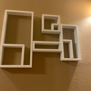 May include: White wall-mounted shelf unit with five rectangular compartments of varying sizes. The shelves are arranged in a geometric pattern.
