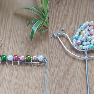 May include: Two wire sculptures of snails decorated with colourful beads. The first snail is a simple design with a single row of beads in shades of pink, green, and white. The second snail has a more elaborate shell design with multiple layers of beads in shades of white, pink, and blue.