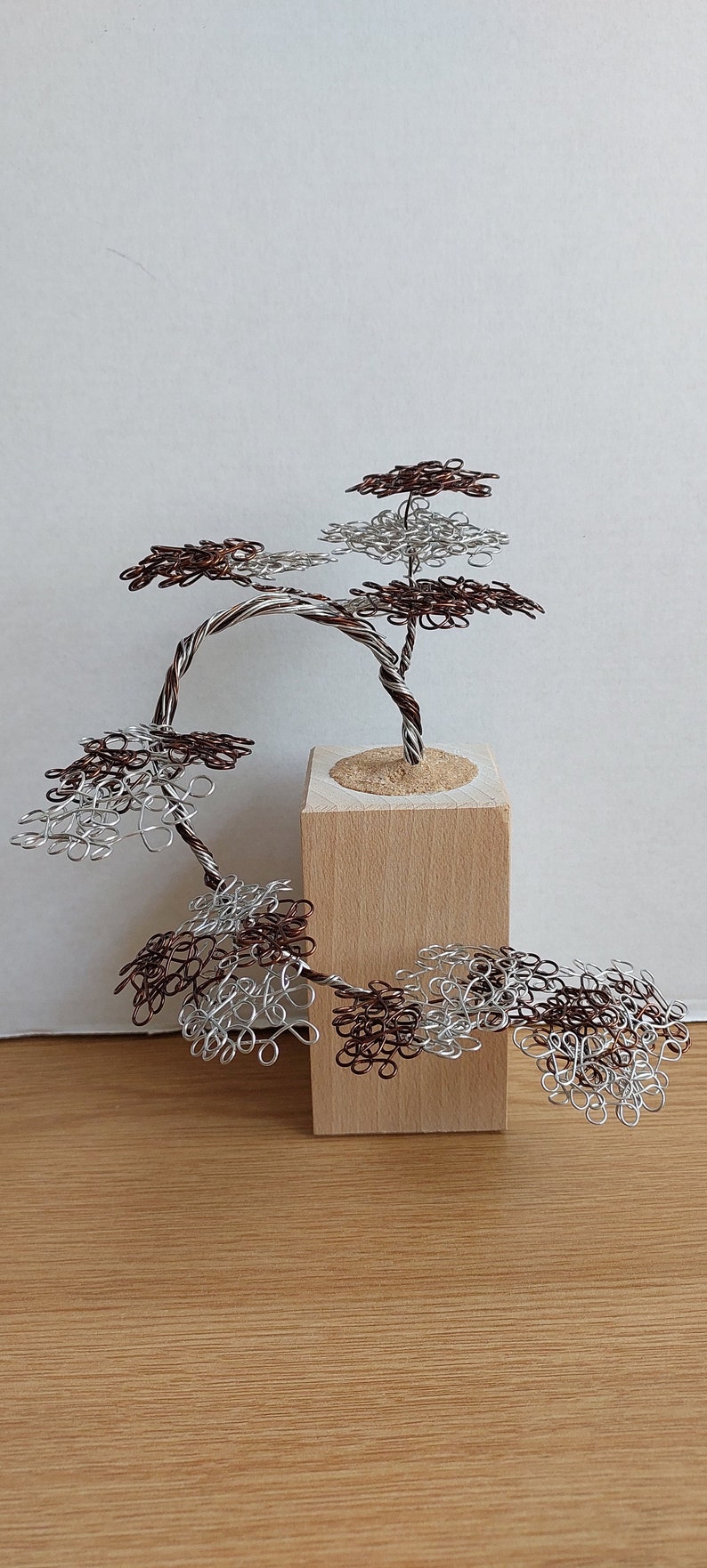 May include: A wire sculpture of a bonsai tree with brown and silver wire branches and leaves. The tree is mounted on a wooden base.