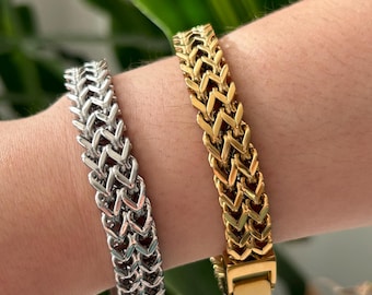 May include: Two metal bracelets, one silver and one gold, are displayed. The bracelets have a woven design, with a series of interlocking chevron shapes. The gold bracelet has a rectangular clasp. The bracelets are shown on a person's wrist.