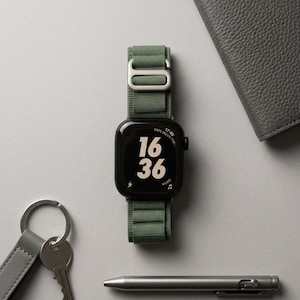 May include: A black smartwatch with a green nylon band displays the time 16:36. The watch is next to a gray leather wallet, a silver pen, and a keychain with keys. The watch band has a silver buckle.