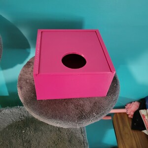 May include: A bright pink square tissue box with a round opening on the top. The box is sitting on a grey fluffy surface.