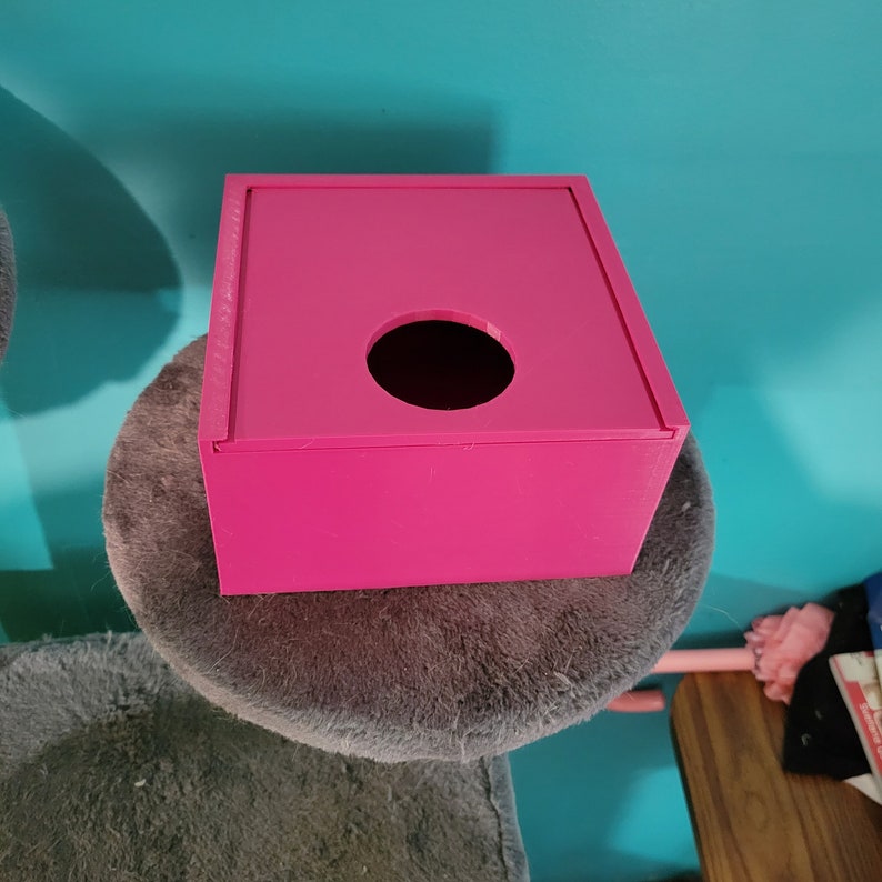May include: A bright pink square tissue box with a round opening on the top. The box is sitting on a grey fluffy surface.