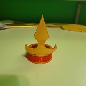May include: A gold 3D printed crown with a red base. The crown has a pointed top and two curved sides.