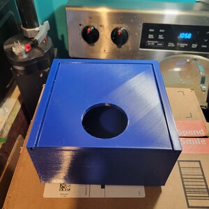 May include: A blue plastic box with a round hole in the top. The box is sitting on a brown cardboard box.
