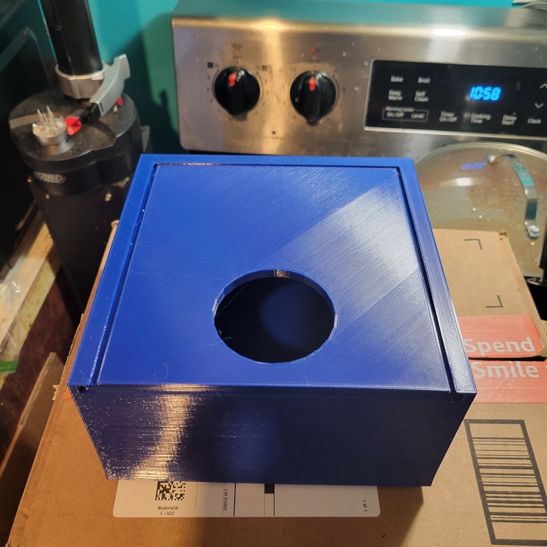 May include: A blue plastic box with a round hole in the top. The box is sitting on a brown cardboard box.