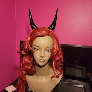 May include: A red wig with black devil horns. The wig is styled in loose curls and the horns are pointed and curved.