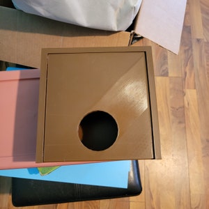 May include: A brown square box with a round hole in the centre. The box has a smooth, slightly shiny surface.