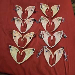 May include: A set of ten pairs of elf ears with various decorative charms and gemstones. The ears are made of a flesh-toned material and are designed to be worn as a costume accessory.