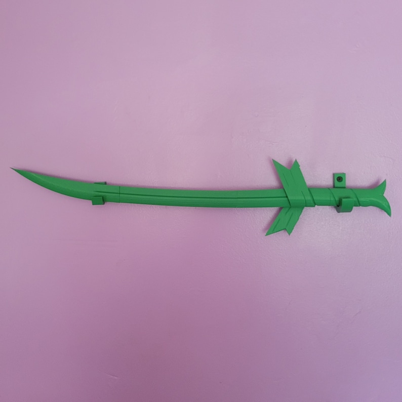 K&ouml;nnte beinhalten: Ein leuchtend gr&uuml;nes, gebogenes Schwert mit einem stilisierten Griff und Parierstange. Die Klinge hat eine Spitze, und der Griff weist dekorative Elemente auf. Das Schwert ist an einer rosa Wand befestigt.