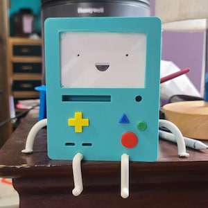 May include: A blue and white 3D printed figurine of a character from the cartoon Adventure Time. The figurine is shaped like a game console with a smiling face and buttons. The figurine has white legs and is sitting on a wooden surface.