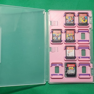 May include: A pink plastic case with clear plastic cover for storing Nintendo Switch game cartridges. The case has eight slots for cartridges and purple accents. The case is filled with various game cartridges.